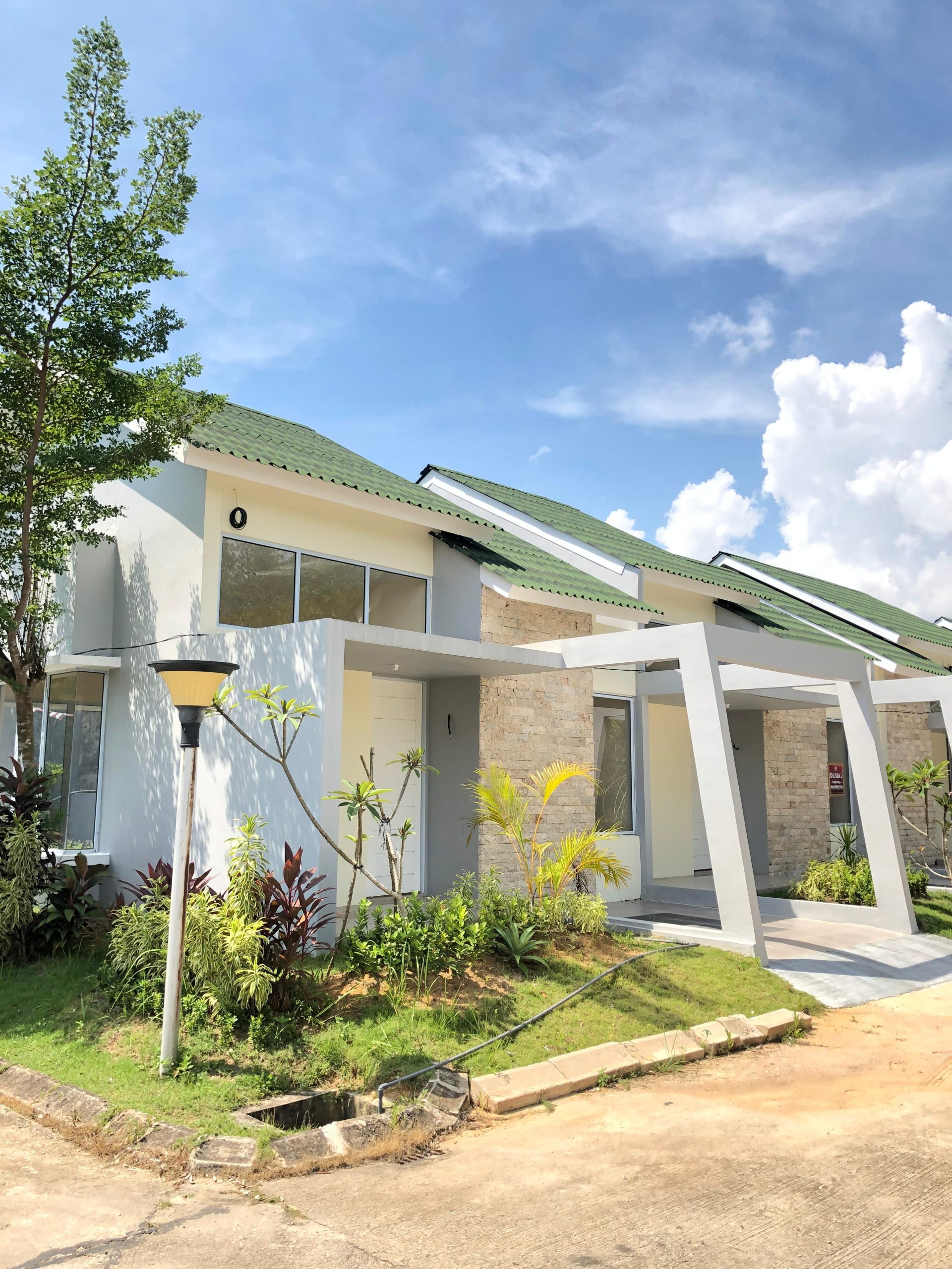 sebuah rumah modern dengan desain minimalis. Bagian depan rumah memiliki teras kecil dengan kolom penopang yang menyatu dengan struktur atap hijau. Di sisi kiri dan kanan teras, terdapat tanaman hijau yang memberikan kesan asri. Dinding rumah terlihat menggunakan bahan batu alam yang memberi nuansa alami, sementara jendela dan pintu utama berdesain sederhana dan bersih. Langit biru cerah dan awan putih yang melatari gambar menambah kesan terang dan segar pada suasana sekitar.